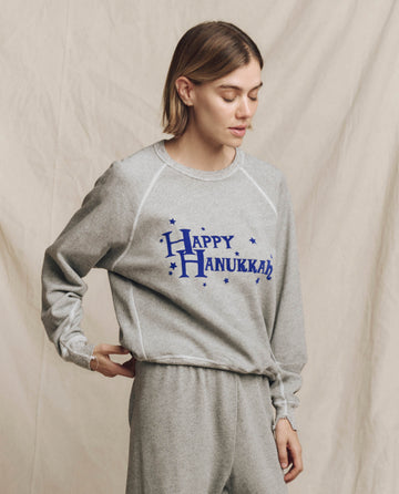 A person wearing "The College Sweatshirt" by THE GREAT. HOLIDAY 24 GRAPHICS in Varsity Grey, featuring a Hanukkah graphic, stands against a plain backdrop, exuding effortless style and cheerful holiday spirit.