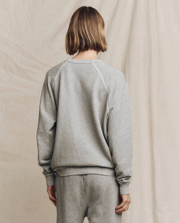 A person with shoulder-length hair is seen wearing The College Sweatshirt in Varsity Grey with a Hanukkah graphic by THE GREAT. HOLIDAY 24 GRAPHICS, along with sweatpants, against a neutral-colored backdrop.