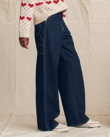 Wearing THE GREAT. PS26 The Mill Jean—Rinse with Cream Stitching, paired with a white sweater with red heart pattern and white sneakers, person stands against a neutral fabric backdrop.