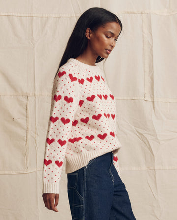 A woman stands against a beige fabric backdrop, wearing THE GREAT. PS26 "The Heart Crew." sweater in Pearl with Red, paired with dark blue jeans.