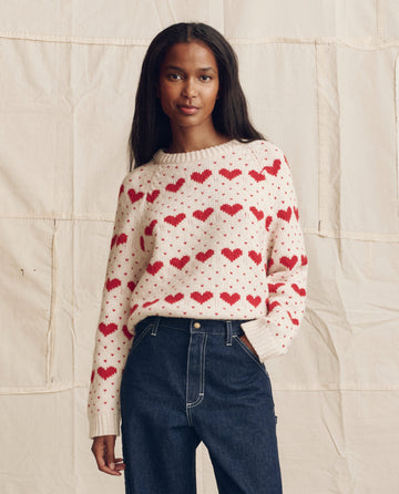 A woman stands before a beige fabric backdrop wearing THE GREAT. PS26's The Heart Crew. sweater in pearl with red and dark blue high-waisted jeans.