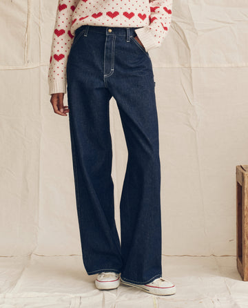Wearing THE GREAT. PS26 The Mill Jean in Rinse with Cream Stitching—high-waisted, wide-leg dark blue cotton-hemp denim—paired with a white sweater with red hearts and white sneakers against a neutral background.