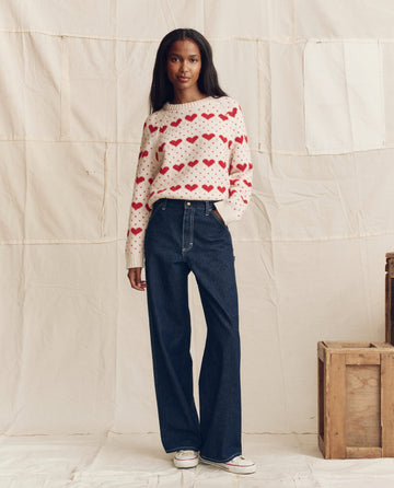 A woman stands indoors wearing a white sweater with red hearts, THE GREAT. PS26 The Mill Jean in Rinse with Cream Stitching, and white sneakers, posed in front of wooden crates and a beige fabric backdrop.