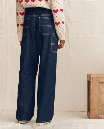 A person faces away, wearing THE GREAT. PS26 The Mill Jean in Rinse with Cream Stitching and a white sweater with red heart patterns. To the right is a wooden crate, all set against a neutral fabric backdrop.