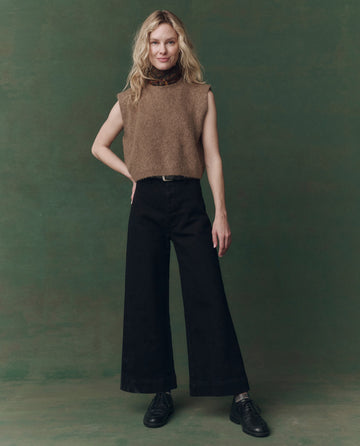 A woman stands against a green backdrop wearing THE GREAT. F25 D2's "The Brushed Melange Sweater – Doe" over a patterned turtleneck, paired with black wide-leg pants and black shoes.