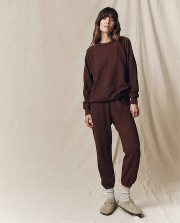 A person stands against a beige fabric backdrop, wearing The Stadium Sweatpant. Solid -- Driftwood from THE GREAT. F25 SEASONAL KNITS, styled with co-ord sweatshirt, gray socks, and beige slip-on shoes—capturing relaxed Los Angeles style.