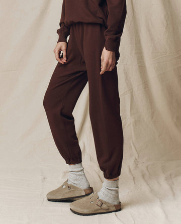A person wears THE GREAT. F25 SEASONAL KNITS The Stadium Sweatpant in Driftwood, paired with a brown long-sleeve top, gray socks, and beige slip-on sandals, standing against a light beige fabric backdrop in Los Angeles.