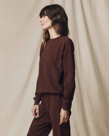 A woman stands in profile before a beige backdrop, wearing THE GREAT.'s The College Sweatshirt in Solid Driftwood from the F25 SEASONAL KNITS collection, styled with matching pants and crafted in Los Angeles.