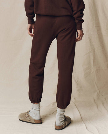 A person stands on a beige fabric surface, wearing THE GREAT. F25 SEASONAL KNITS The Stadium Sweatpant in Driftwood with matching sweatshirt, gray socks, and beige slip-on sandals. The image shows a relaxed lower body from behind.