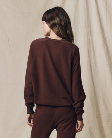Person stands with back to camera, wearing The College Sweatshirt in Driftwood from THE GREAT. F25 SEASONAL KNITS, paired with matching pants, set against a beige fabric backdrop.