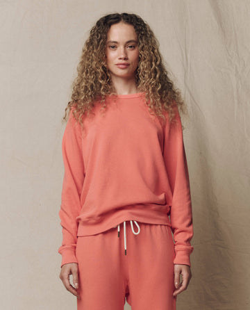 A woman with curly hair wears THE GREAT. SU26 SEASONAL CORE The College Sweatshirt in Solid Guava, standing against a neutral fabric backdrop and facing the camera.