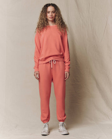 A person stands facing forward, wearing THE GREAT. SU26 SEASONAL CORE's The College Sweatshirt in Solid Guava, paired with matching sweatpants and white high-top sneakers, in front of a beige fabric backdrop.