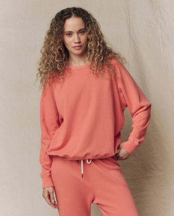 A woman with curly hair wears THE GREAT. SU26 SEASONAL CORE The College Sweatshirt in Solid Guava, paired with matching sweatpants, standing against a neutral fabric backdrop.