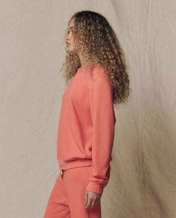 A person with long curly hair wears THE GREAT. SU26 SEASONAL CORE The College Sweatshirt, Solid -- Guava, and matching pants in profile against a beige fabric background.