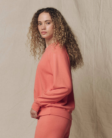 A person with curly hair wears THE GREAT. SU26 SEASONAL CORE The College Sweatshirt. Solid -- Guava, standing against a neutral fabric background and looking at the camera.