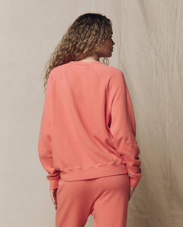 Person with curly hair wears THE GREAT. SU26 SEASONAL CORE "The College Sweatshirt. Solid -- Guava" and matching pants, standing with their back to the camera against a neutral fabric backdrop.