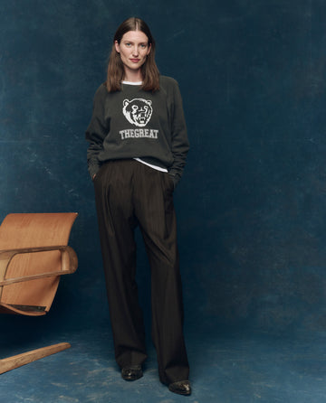 A woman stands with hands in pockets, wearing a dark bear graphic sweatshirt and THE GREAT. F25 COLLECTORS The Menswear Trouser. -- Chocolate Stripe, paired with black shoes beside a wooden chair against a blue backdrop.