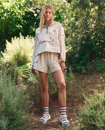 Outdoors among greenery, a person wears THE GREAT. SP26 TGO’s Embroidered Cropped Trailhead Pullover in cream with black daisies, paired with shorts, striped socks, and white sandals.