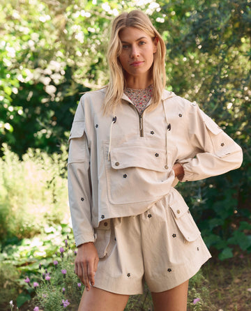Wearing THE GREAT. SP26 TGO's Embroidered Cropped Trailhead Pullover in cream with black daisies, a person stands at a trailhead surrounded by greenery.