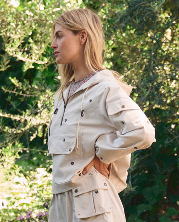A woman with blonde hair stands outdoors in THE GREAT. SP26 TGO’s Embroidered Cropped Trailhead Pullover in Cream with Black Daisies, paired with matching pants; green foliage fills the background.