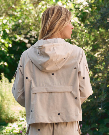 A woman with blonde hair stands outdoors amid lush greenery, wearing THE GREAT. SP26 TGO's Embroidered Cropped Trailhead Pullover in cream with black daisies, facing away from the camera.