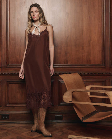 A woman in THE GREAT. F25 D1 The Legacy Dress in Chocolate, embroidered with mesh lace, paired with tan cowboy boots and a white neck scarf, stands by a wooden chair against a paneled wood wall.