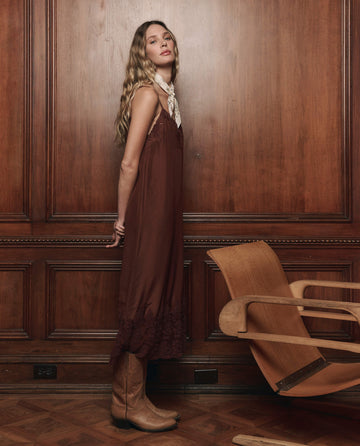 A woman wearing THE GREAT. F25 D1's The Legacy Dress in Chocolate stands beside a wooden chair against a wood-paneled wall, gazing at the camera, her look evocative of 1920s loungewear and finished with cowboy boots.