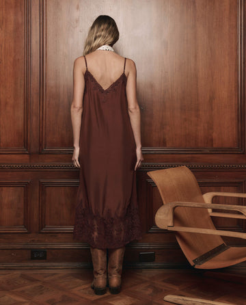 A woman with long hair stands facing a wood-paneled wall, wearing THE GREAT. F25 D1 The Legacy Dress -- Chocolate and boots. An empty wooden chair is positioned nearby on a parquet floor.