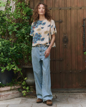 A person stands outdoors in front of a wooden door wearing THE GREAT. PF25 The Mandolin Top in Sungold Floral, paired with loose light blue jeans and brown shoes, surrounded by green plants.