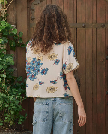 A person with long curly hair wears THE GREAT. PF25's The Mandolin Top in Sungold Floral and light blue jeans, standing by a wooden door framed with lush greenery.
