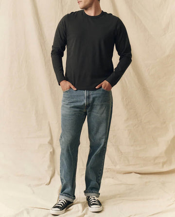 A person stands with hands in pockets, wearing THE GREAT. FALL 23 MEN’s The Men's Pure Knits Long Sleeve Slim Crew in Solid -- Almost Black, blue jeans, and black-and-white sneakers against a beige fabric backdrop.