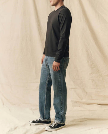 A person stands sideways wearing THE GREAT. FALL 23 MEN The Men's Pure Knits Long Sleeve Slim Crew in Solid -- Almost Black, paired with blue jeans and black sneakers, set against a beige fabric backdrop.