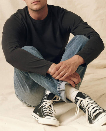 A person sits cross-legged, wearing THE GREAT. FALL 23 MEN's The Men's Pure Knits Long Sleeve Slim Crew in Almost Black, paired with light blue jeans, white socks, and black high-top sneakers.