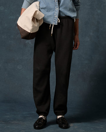 A person stands against a dark, textured background wearing a light denim shirt with THE GREAT. F26 D1 FLEECE BACK KNITS The Fleece Stadium Sweatpant in Solid — Almost Black, black shoes, and holding a two-tone bag.