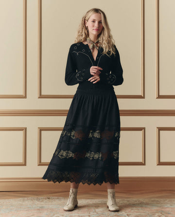 A woman stands indoors against a paneled wall, wearing THE GREAT. SP26 D2's black Rosette Crochet Canary Skirt, a black embroidered blouse, a scarf around her neck, and light-colored ankle boots.