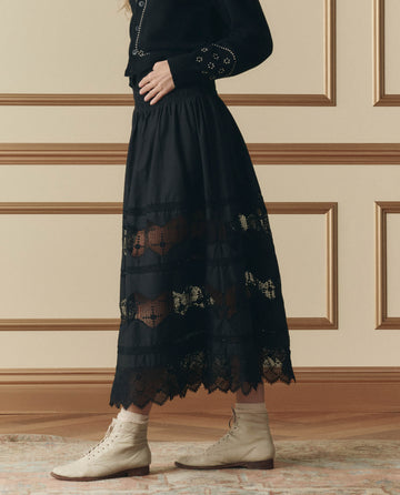 A person wearing THE GREAT. SP26 D2’s The Rosette Crochet Canary Skirt in black, paired with a black sweater and beige lace-up ankle boots, stands indoors on a patterned rug.