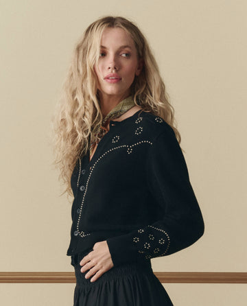 A woman with long, wavy blonde hair wears THE GREAT. SP26 D2 The Studded Cardigan in black, paired with a black skirt and light scarf, standing against a beige background.