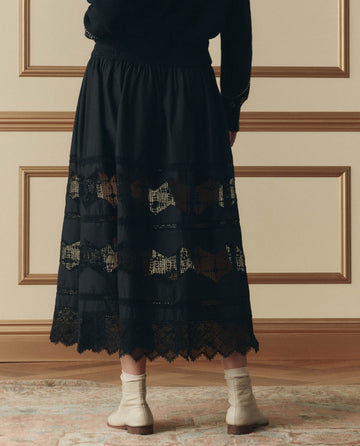 Person indoors, shown from the back against paneled walls, wears THE GREAT. SP26 D2 The Rosette Crochet Canary Skirt in black, styled with cream socks and beige shoes.