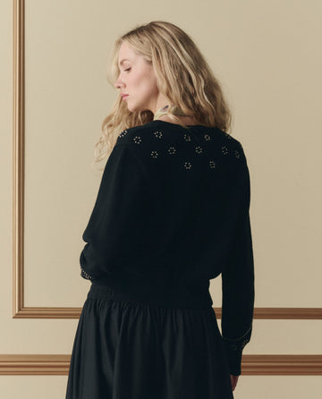 A woman with long blonde hair, wearing THE GREAT. SP26 D2 Studded Cardigan in black, stands facing away against a beige wall with decorative molding.
