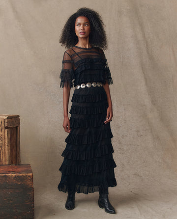 A woman models THE GREAT. H25 COLLECTION's The Winter Mesh Flounce Top in black, styled with tiered ruffle dress, wide silver belt, and black boots, against a neutral fabric backdrop with wooden boxes.