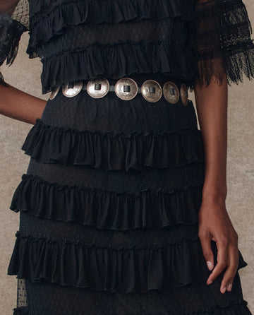 A person wears THE GREAT. H25 COLLECTION's The Winter Mesh Flounce Skirt in black, styled with sheer sleeves and a wide metallic belt adorned with large circular discs.