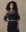 A woman with curly hair models THE GREAT. H25 COLLECTION's Winter Mesh Flounce Top in black, featuring sheer ruffles and layered details, styled with a metallic belt against a neutral background.