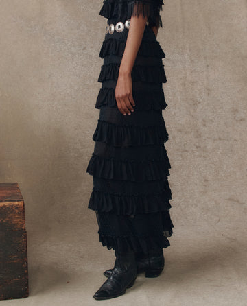 A person wearing THE GREAT. H25 COLLECTION's The Winter Mesh Flounce Skirt in black, paired with a black top, silver concho belt, and black pointed-toe boots, stands against a neutral backdrop.