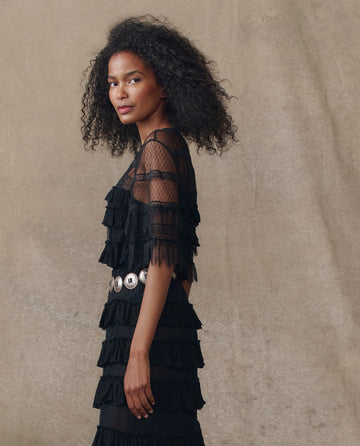 A woman with curly hair models THE GREAT. H25 COLLECTION’s The Winter Mesh Flounce Top in black, styled with a silver belt against a beige background—a chic winter fashion statement.