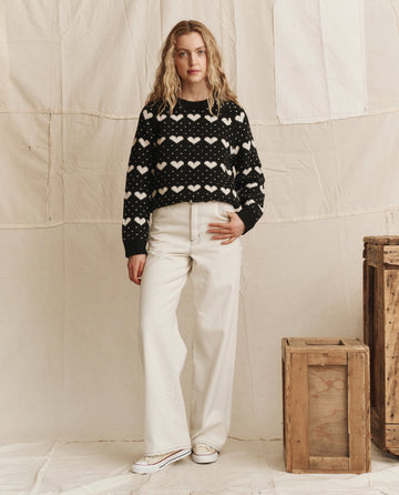 A woman stands indoors next to wooden crates against a neutral backdrop, wearing THE GREAT. PS26's The Heart Crew. in Black With Pearl, off-white wide-leg pants, and white sneakers.