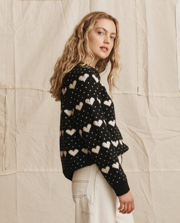 A woman with wavy blonde hair stands in profile against a beige fabric backdrop, wearing THE GREAT. PS26’s The Heart Crew. sweater in Black With Pearl, paired with white pants.