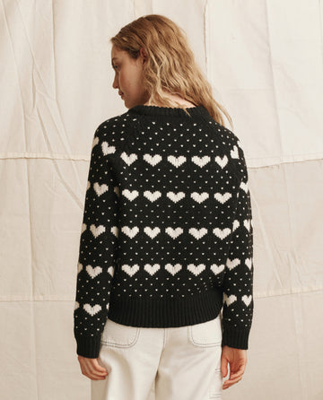 A person with wavy hair stands against a neutral backdrop wearing THE GREAT. PS26's The Heart Crew.—a black sweater with rows of white hearts and small dots—paired with light-colored pants.
