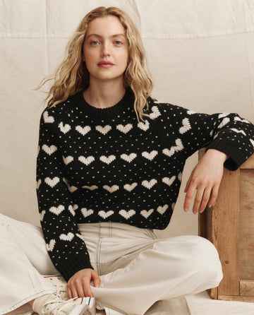 A person with wavy blonde hair sits cross-legged against a neutral background, wearing THE GREAT. PS26 The Heart Crew. -- Black With Pearl sweater and white pants.