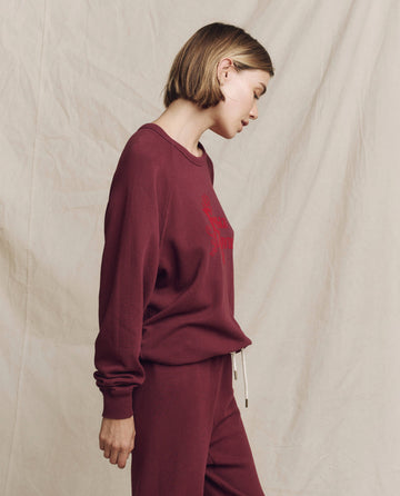 A person wearing The College Sweatshirt in mulled wine featuring a Feliz Navidad graphic, along with matching pants, stands against a beige fabric background.