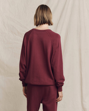 A person in a maroon "The College Sweatshirt" with a Feliz Navidad graphic is seen from behind against a light-colored fabric backdrop, wearing the vintage-style attire from THE GREAT. HOLIDAY 24 GRAPHICS collection.